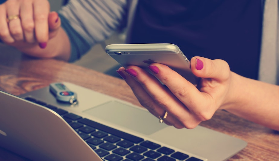 A woman looks at a mobile phone while at a laptop computer. by Firmbee.com from Unsplash